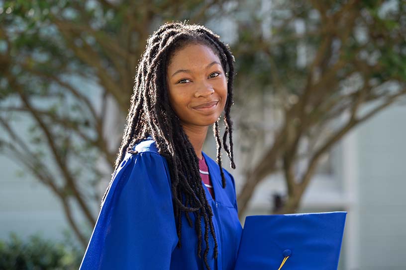 A young woman facing the camera, wearing a blue graduation gown while holding a blue graduation cap.