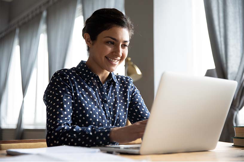 A woman sitting at a table, smiling while looking at a laptop screen.